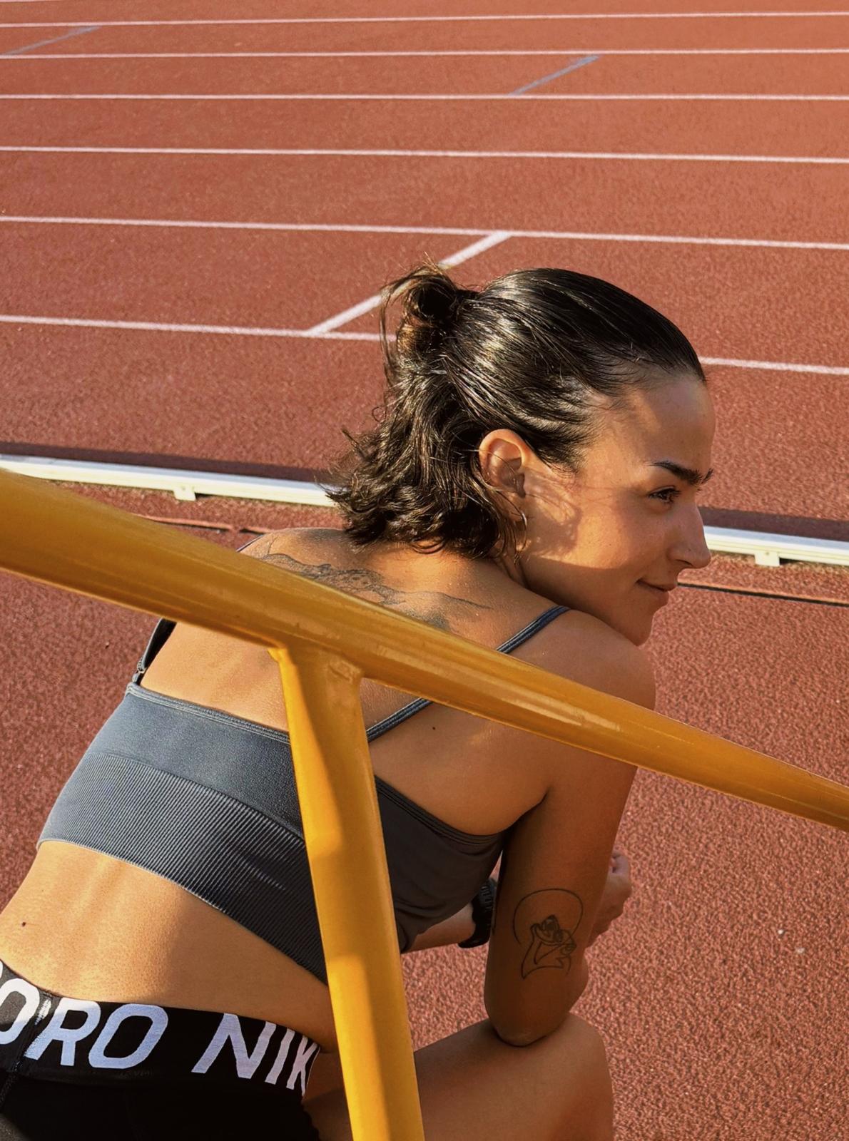 Maria resting on a yellow railing at the athletics track, looking back over her shoulder